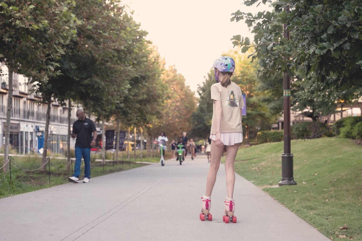A kid in a helmet and roller skates on a wide, tree-lined section of the Beltline, scooters and walkers ahead